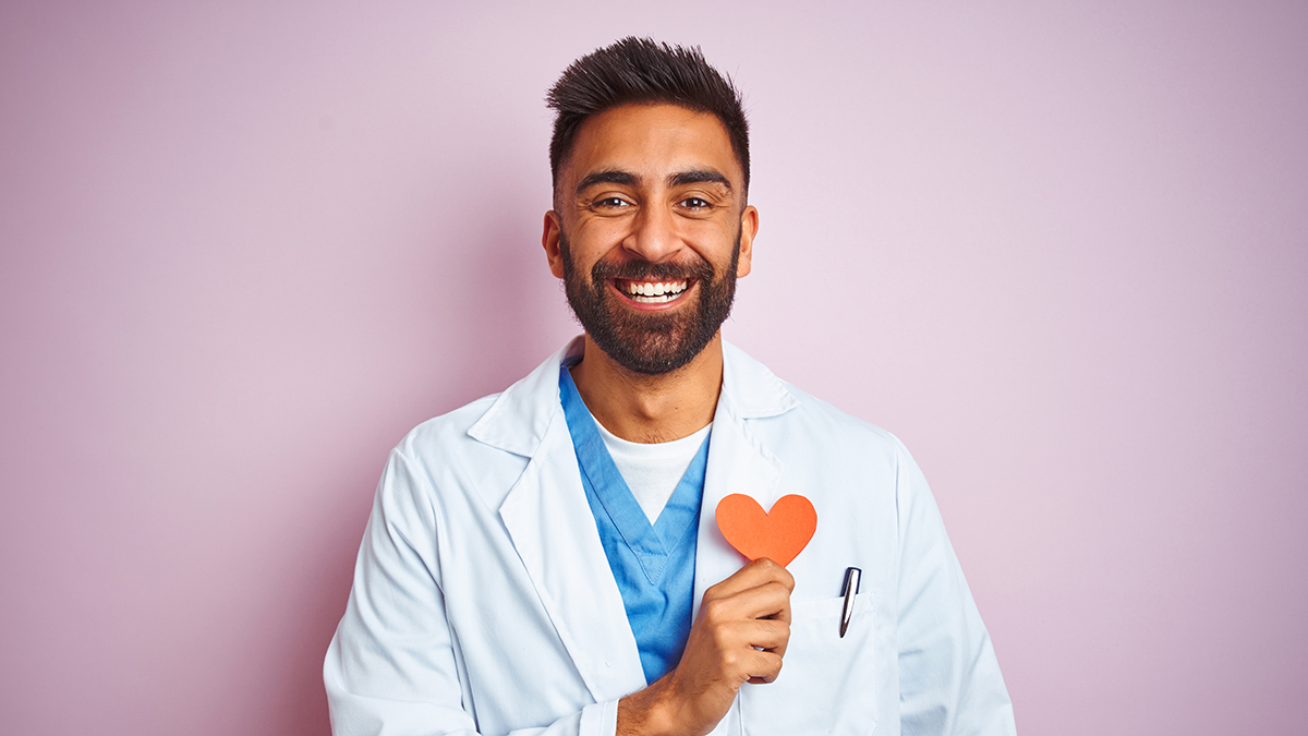 Young smiling doctor holding paper heart to chest.